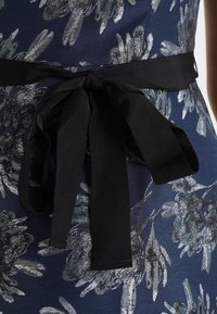 Tkanina w kwiatowy wzór w odcieniach granatu i srebra z czarnym paskiem zawiązanym w kokardę, ukazująca gładką strukturę i akcenty kwiatowe.