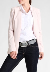 Helles rosafarbenes Blazer über einem weißen Hemd mit Knöpfen, kombiniert mit dunklen Jeans und einem grauen Gürtel, der ein schlichtes Design und eine glatte Textur aufweist.