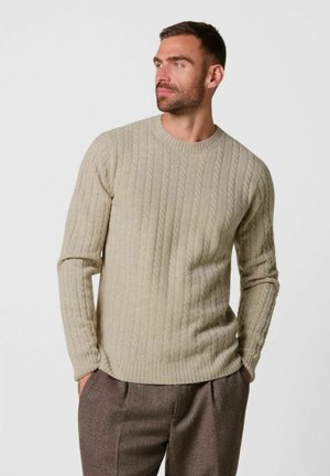 Hombre que lleva un jersey beige de punto trenzado y pantalones marrones, de pie con las manos en los bolsillos, mirando hacia un lado con un fondo liso.