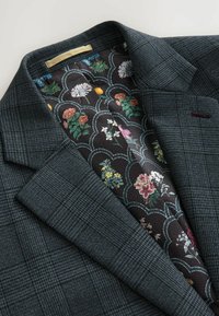 Sacou în carouri de culoare verde închis, cu o căptușeală neagră cu model floral. Are rever cu colțuri și o broderie subtilă de culoare burgund.