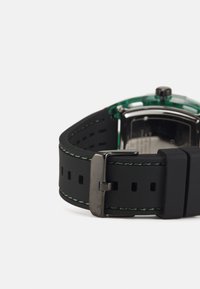 Correa de reloj de goma negra con costuras verdes, hebilla de acero inoxidable y un fondo de caja negro visible. Forma redonda distintiva con elementos transparentes.