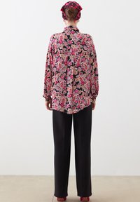 Long-sleeve floral blouse in pink and black, featuring a vibrant paisley pattern, button-down back, paired with black trousers and pink heels.