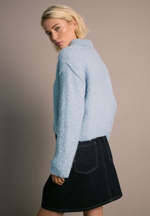 Giovane donna con capelli biondi corti che indossa un maglione azzurro chiaro soffice e una gonna in denim nera, guarda oltre la spalla.