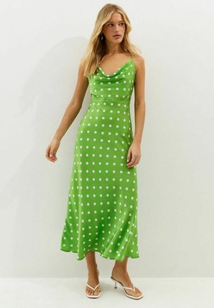 Femme portant une robe midi verte sans manches à pois blancs et des sandales à talons ouvertes blanches, se tenant devant un fond uni.