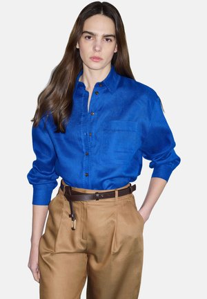 Frau mit langen braunen Haaren, die ein leuchtend blaues Hemd mit Knöpfen trägt, das in hoch taillierte beige Hosen mit einem dunkelbraunen Gürtel gesteckt ist.