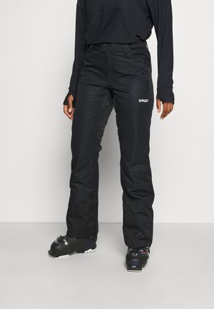 Personne porte un pantalon de neige Oakley noir avec un haut noir et des chaussures de ski noires, debout sur un fond gris clair uni.