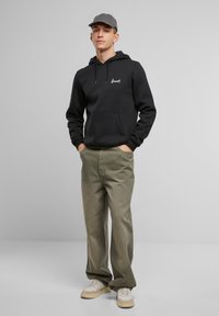 Felpa nera con tasca frontale, logo bianco, pantaloni verde oliva e scarpe leggere, completati da un cappellino grigio, in piedi contro uno sfondo neutro.