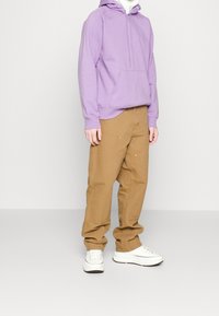 Felpa viola con tasca frontale, abbinata a pantaloni beige larghi con cuciture visibili e accenti sottili. Sneakers bianche.