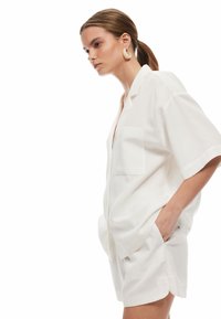 Camicia bianca a maniche corte con tasca, abbinata a pantaloni corti coordinati. Tessuto liscio e leggero, vestibilità comoda con una leggera lucentezza.