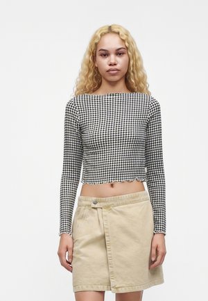 Mladá žena s dlhými kučeravými blond vlasmi, ktorá nosí čierno-biely kockovaný dlhý rukáv crop top a béžovú sukňu na jednoduchom pozadí.
