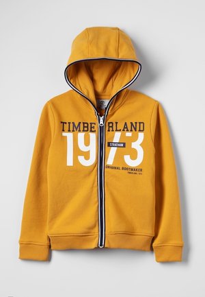 Gul hættetrøje med lynlås, sort og hvid kant, hætte og teksten "TIMBERLAND 1973 Original Bootmaker Stratham" på forsiden.