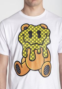 Camiseta blanca con un gráfico de un oso de dibujos animados. El oso es amarillo con un diseño derretido, contorneado en negro, y tiene un patrón de cuadros en su cuerpo.