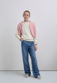 Hanorac bej deschis cu mâneci bufante, pulover roz purtat pe umeri și blugi albaștri cu picioare evazate. Încălțămintea are un design cu model.