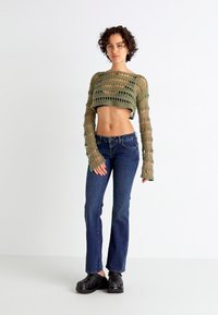Heklet grønn bustetopp med lange ermer, kombinert med mørkeblå utsvingte jeans og svarte tresko. Toppen har et åpent, strukturert mønster.