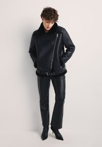 Czarna skórzana kurtka z kołnierzem z owczej wełny, z ukośnym zamkiem błyskawicznym i ściągniętym w pasie krojem. Połączona z dopasowanymi czarnymi skórzanymi spodniami i szpilkami.