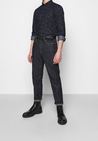 Camicia a bottone a righe navy con maniche arrotolate, jeans in denim scuro e stivali neri con lacci. Cintura con fibbia in metallo in evidenza.