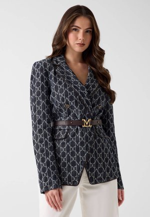 Femme portant un blazer foncé à motifs avec des boutons dorés et une ceinture marron ornée d'une boucle "M" en or, associée à un pantalon de couleur claire.