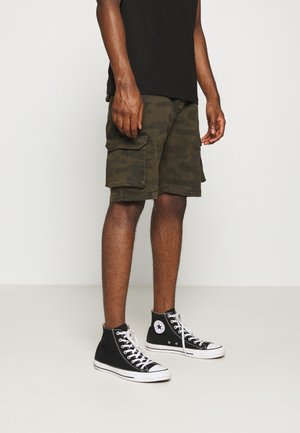 Man in zwarte high-top Converse sneakers, camouflage cargo shorts en een zwart t-shirt staat op een effen vloer tegen een witte muur.