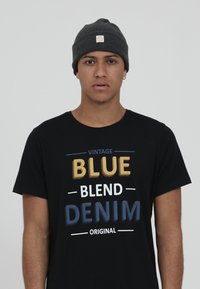 Black t-shirt with colorful text: "VINTAGE BLUE BLEND DENIM ORIGINAL." Grey beanie hat. Model has short hair and an earring.