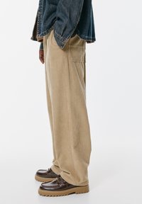 Bežové kordové nohavice s uvoľneným strihom, so bočnými vreckami. Skombinované s tmavohnedými topánkami, ktoré majú textúrovaný povrch a masívnu podrážku.