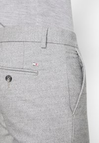 Närbild av ljusgrå texturerade byxor med bältesloop, sidofickor och bakfickor, samt en liten rektangulär logotyp i röd, vit och svart.