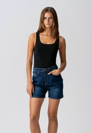 Frau steht vor einem schlichten Hintergrund, trägt ein schwarzes ärmelloses Oberteil und dunkelblaue Jeansshorts, eine Hand in der Tasche.