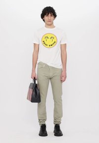 Valge T-särk, millel on ümmargune graafiline disain palmi puudega ja tekst "PAUL SMITH". Oliivrohelised püksid ja must kott triibuliste detailidega.