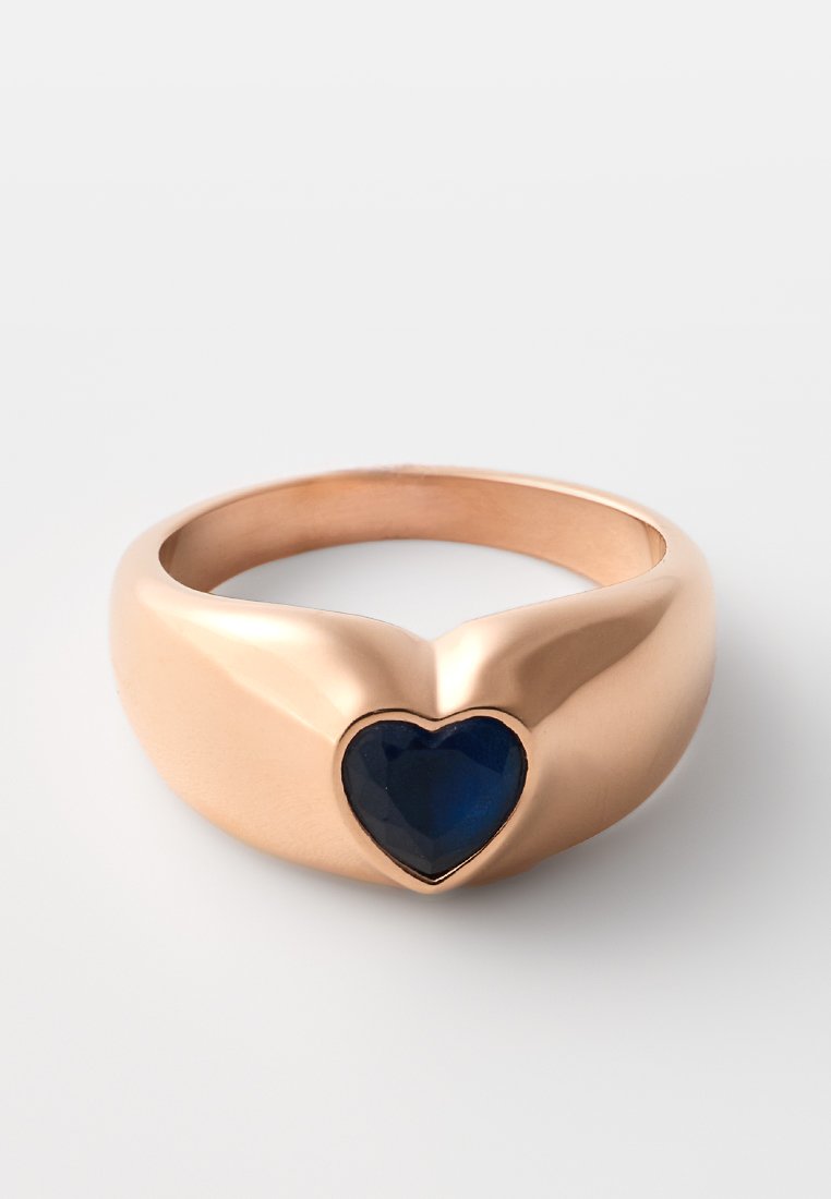 Anillo de oro rosa con una piedra facetada en forma de corazón de color azul oscuro en el centro. Superficie lisa, banda ligeramente cónica con un diseño moderno.