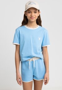 Jasnoniebieska bawełniana koszulka z białymi wykończeniami, z małym logo w kształcie kwiatka. W połączeniu z pasującymi szortami i beżową czapką.