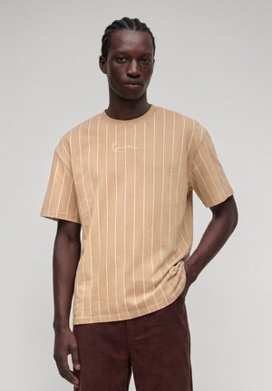Ung mand med flettet hår iført en beige T-shirt med hvide lodrette striber og brune bukser, stående mod en ensfarvet lys baggrund.