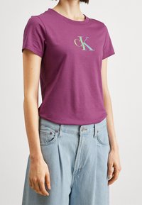Fialové bavlněné tričko s kulatým výstřihem a krátkými rukávy. Na přední straně se nachází světlé logo „cK“. Kombinováno se světle modrými džíny.