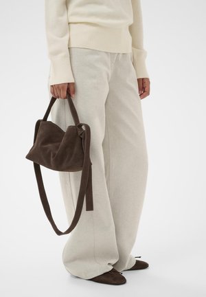 Braune Wildlederhandtasche mit weicher, lässiger Form und langem Riemen, gehalten von einer Person in hellgrauer, weit geschnittener Hose und cremefarbenem Pullover.