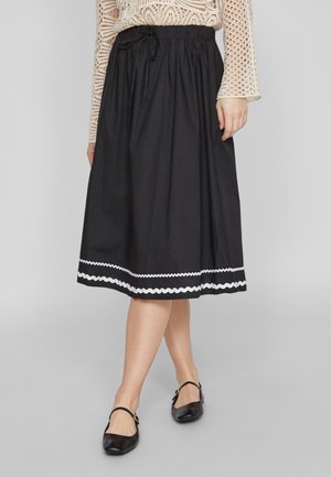 Jupe noire jusqu'aux genoux avec une taille élastique et un cordon de serrage. Équipée d'une bordure blanche en zigzag à l'ourlet. Portée avec des ballerines noires.