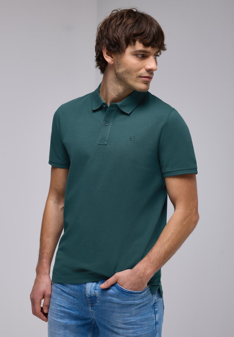 Mörkgrön pikétröja med klassisk krage, tvåknappsslutning och korta ärmar, tillsammans med ljusblå denimjeans.