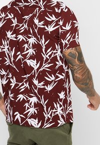 Camicia a maniche corte realizzata in tessuto color marrone scuro con un motivo di foglie di bambù bianche. Presenta un colletto classico e una vestibilità comoda.