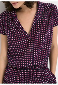 Blusa de manga corta en tejido azul marino oscuro con un patrón de corazones en rosa y blanco. Cuenta con un cuello con solapa y diseño de botones en la parte delantera.