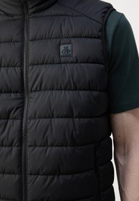 Sort puffervest med quiltet mønster, udstyret med lynlås og et struktureret logo-patch på brystet. Båret over en grøn skjorte.