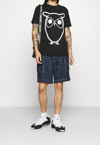 Camiseta de algodón negra con un gráfico de búho blanco, combinada con pantalones cortos de mezclilla con bolsillos cargo y cordón. Zapatillas en blanco y negro.
