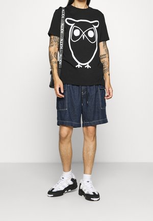 Camiseta de algodón negra con un gráfico de búho blanco, combinada con pantalones cortos de mezclilla con bolsillos cargo y cordón. Zapatillas en blanco y negro.