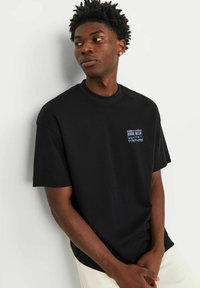 Sort bomulds t-shirt med korte ærmer og rund hals. Har et lille grafisk motiv i lilla og blå på venstre brystområde.