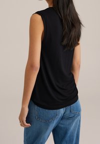 Kvinna med långt mörkt hår som bär en svart ärmlös topp och blå jeans, stående mot en enfärgad beige bakgrund, bakifrån.