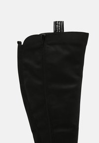 Černé kozačky po kolena vyrobené z hladkého materiálu, s postranním zipem a textilním poutkem se textem. Jednoduchý, elegantní design.