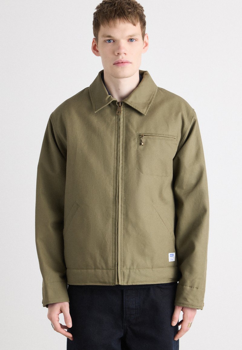 Veste olive verte avec une fermeture éclair à l'avant, un col cranté et une poche poitrine. Fabriquée en matériau durable, elle présente une coupe décontractée et des détails de couture.