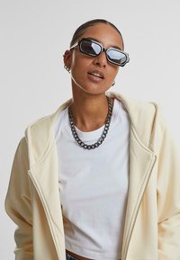 Zonnebril met zwarte hexagonale montuur, een lichtcrème zip-up hoodie en een zilveren ketting om de hals gedragen over een wit T-shirt.