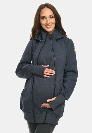 Schwangere Frau lächelt, trägt einen dunkelblauen Umstands-Hoodie mit Frontreißverschlüssen und Taschen und hält ihren Bauch behutsam.