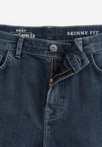 Sötétkék, skinny fit farmernadrág, cipzárral és fém gombos záródással, látható varrási részletekkel. Öt zsebes dizájnnal rendelkezik.