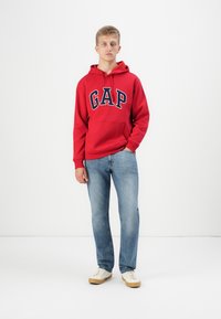 Rode hoodie met "GAP" logo op de voorkant, voorzak, gecombineerd met lichtblauwe jeans en beige sneakers. Glad materiaal, casual pasvorm.