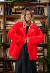 Piros műszőrme kabát cipzáras lezárással, széles gallérral és túlméretezett ujjakkal, fekete ruha fölött viselve. A háttérben könyvespolcok és zöld növények láthatók.