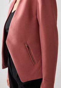 Blazer de couleur framboise en tissu structuré avec une coupe ajustée, équipé d'une poche zippée élégante et d'une coupe minimaliste.