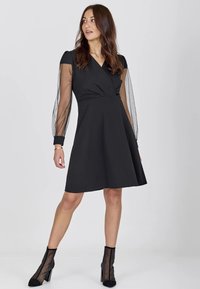 Robe noire avec un corsage ajusté, une jupe évasée et des manches bouffantes en organza transparent, associée à des bottines noires en mesh.